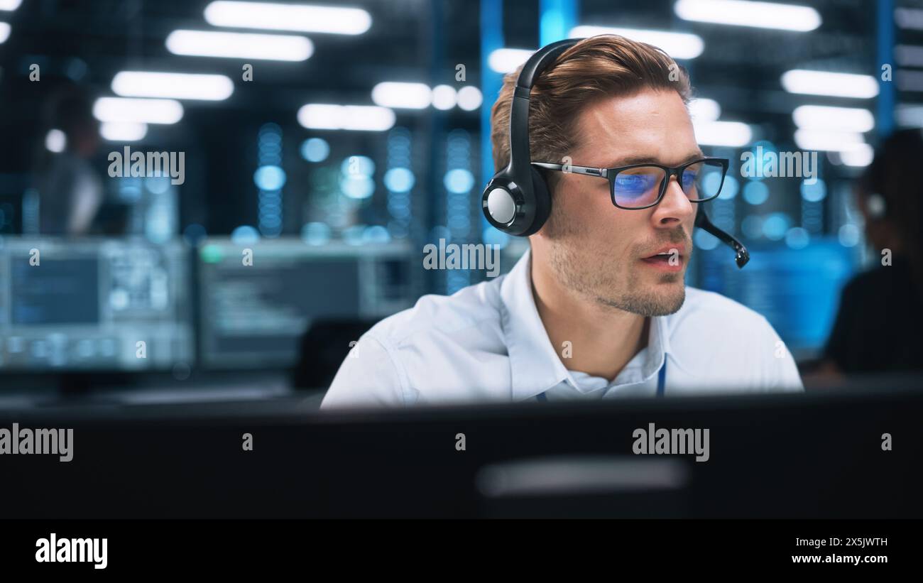 Porträt eines jungen kaukasischen Callcenter-Mitarbeiters, der ein Telefon-Headset trägt und nachts mit ernstem Gesicht in einem modernen Open-Plan-Büro spricht. Helpline-Konzept Stockfoto