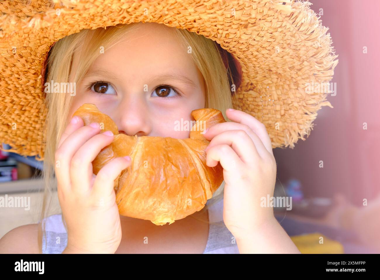 Nahaufnahme des Gesichts des Kindes, Mädchen 3 Jahre alt hält rosa gebackenes Croissant mit Händen nahe dem Mund, zeigt fröhliches Lächeln, Konzept köstliches Frühstück, happ Stockfoto