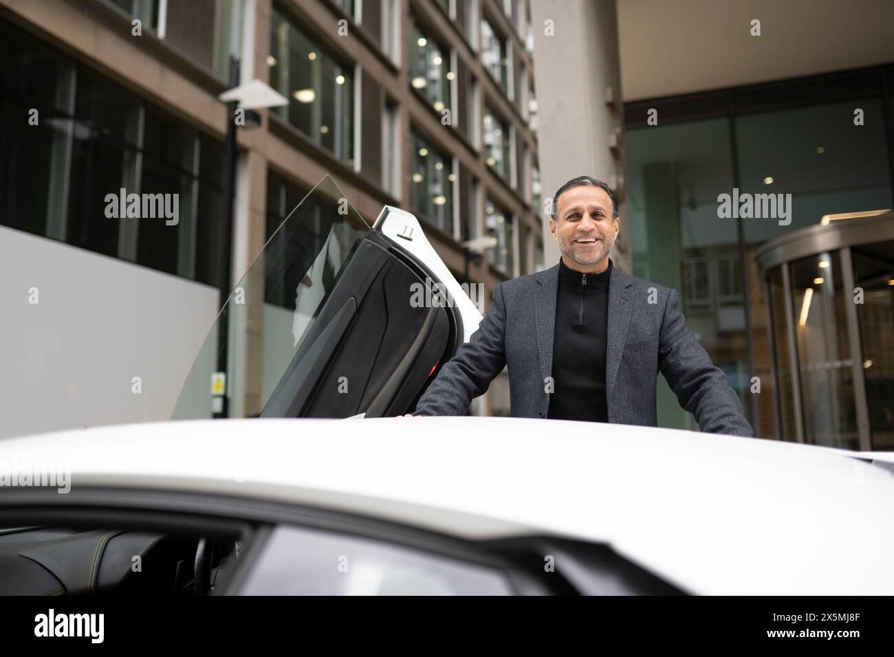 Ein lächelnder reifer Mann, der sich an ein Luxusauto lehnt Stockfoto