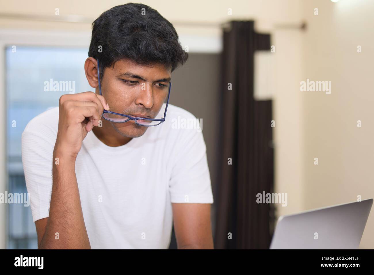 Ein konzentrierter junger Indianer mit Brille arbeitet fleißig an seinem Laptop in seinem Heimbüro. Stockfoto