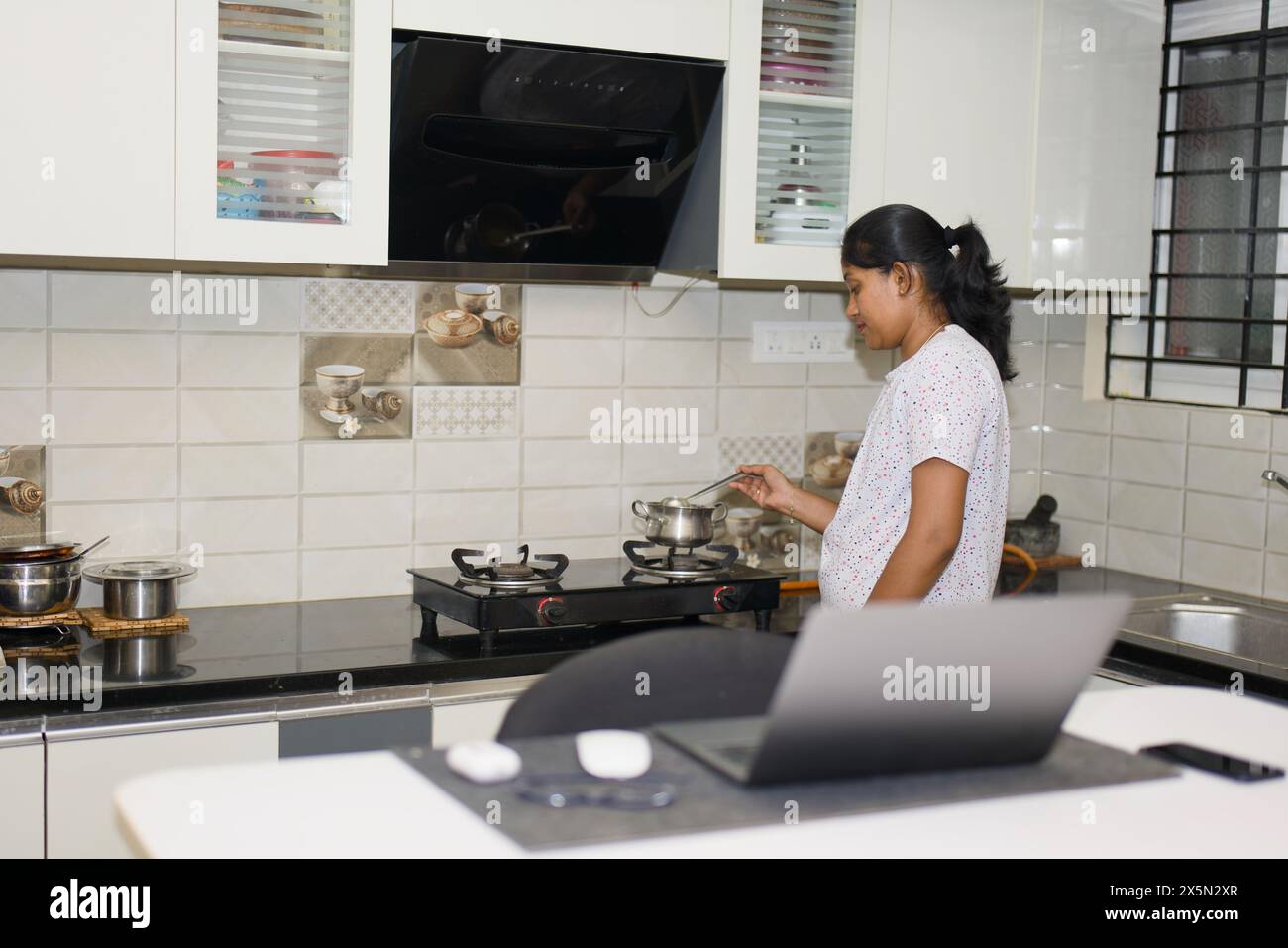 Eine fokussierte junge Frau kocht effizient eine Mahlzeit in ihrer Küche, während sie auf ein Rezept oder eine Anleitung auf ihrem Laptop verweist. Stift Funke Stockfoto