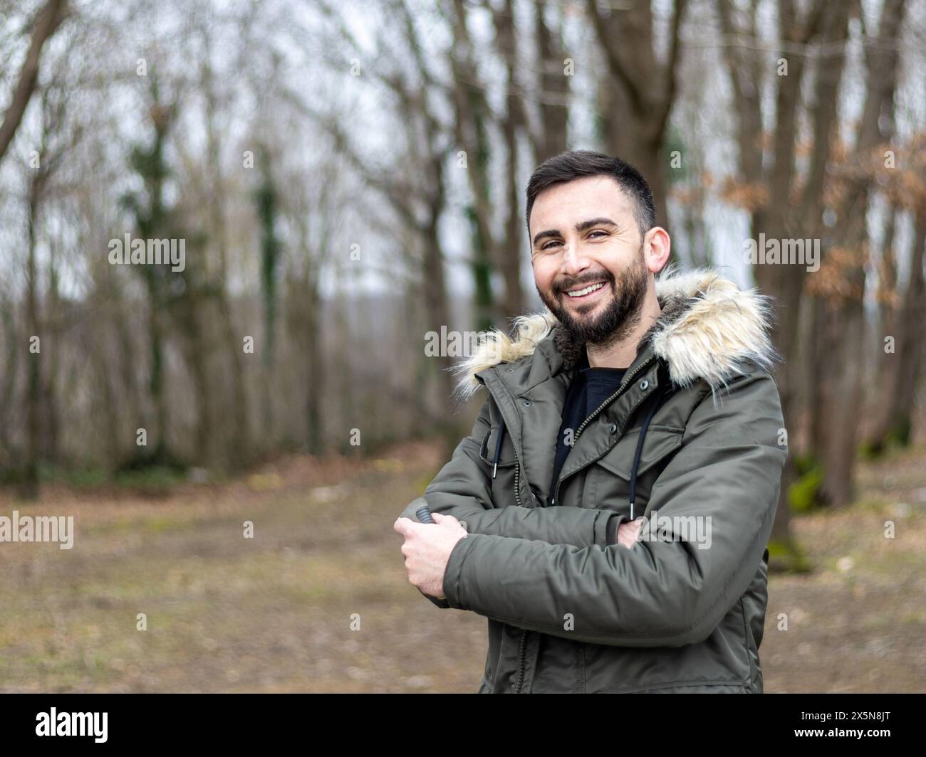 Junger und gutaussehender bärtiger Mann, der in die Kamera schaut und im Park, im Wald lächelt. Ein Mann, der bei kaltem Wetter einen Mantel trägt Stockfoto