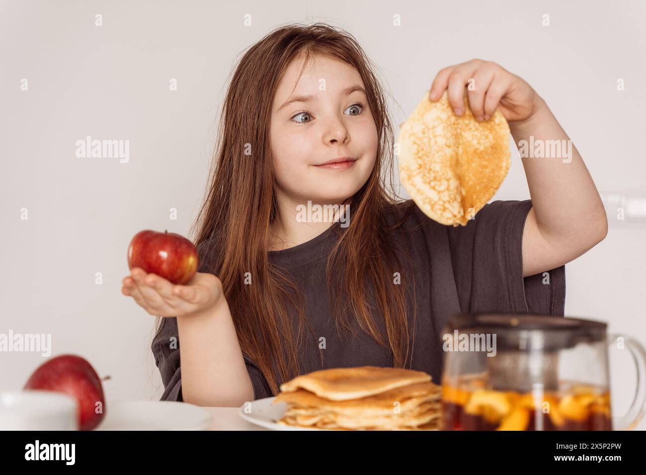 Ein kleines Teenager-Mädchen, das einen Apfel in der einen Hand in der anderen Pfannkuchen hält. Das Mädchen wählt zwischen einem Apfel und einem Pfannkuchen zum Frühstück, sieht aus, als ob es ihn leckt Stockfoto