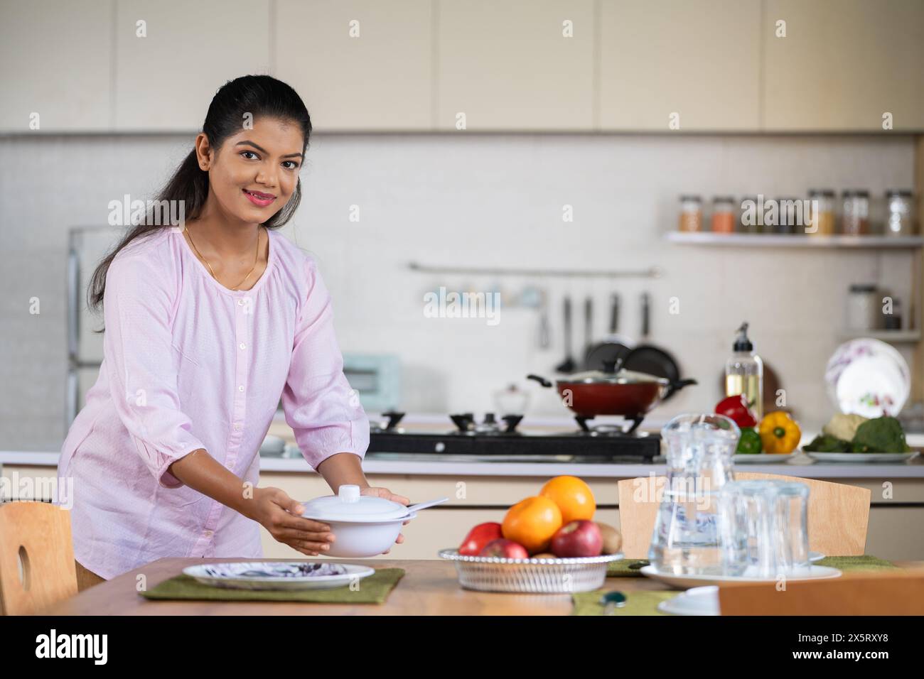 Glückliche indische Frau, die morgens den Esstisch für das Frühstück vorbereitet, während sie die Kamera ansieht - Konzept, tägliche Routine, Familienverantwortung und Stockfoto