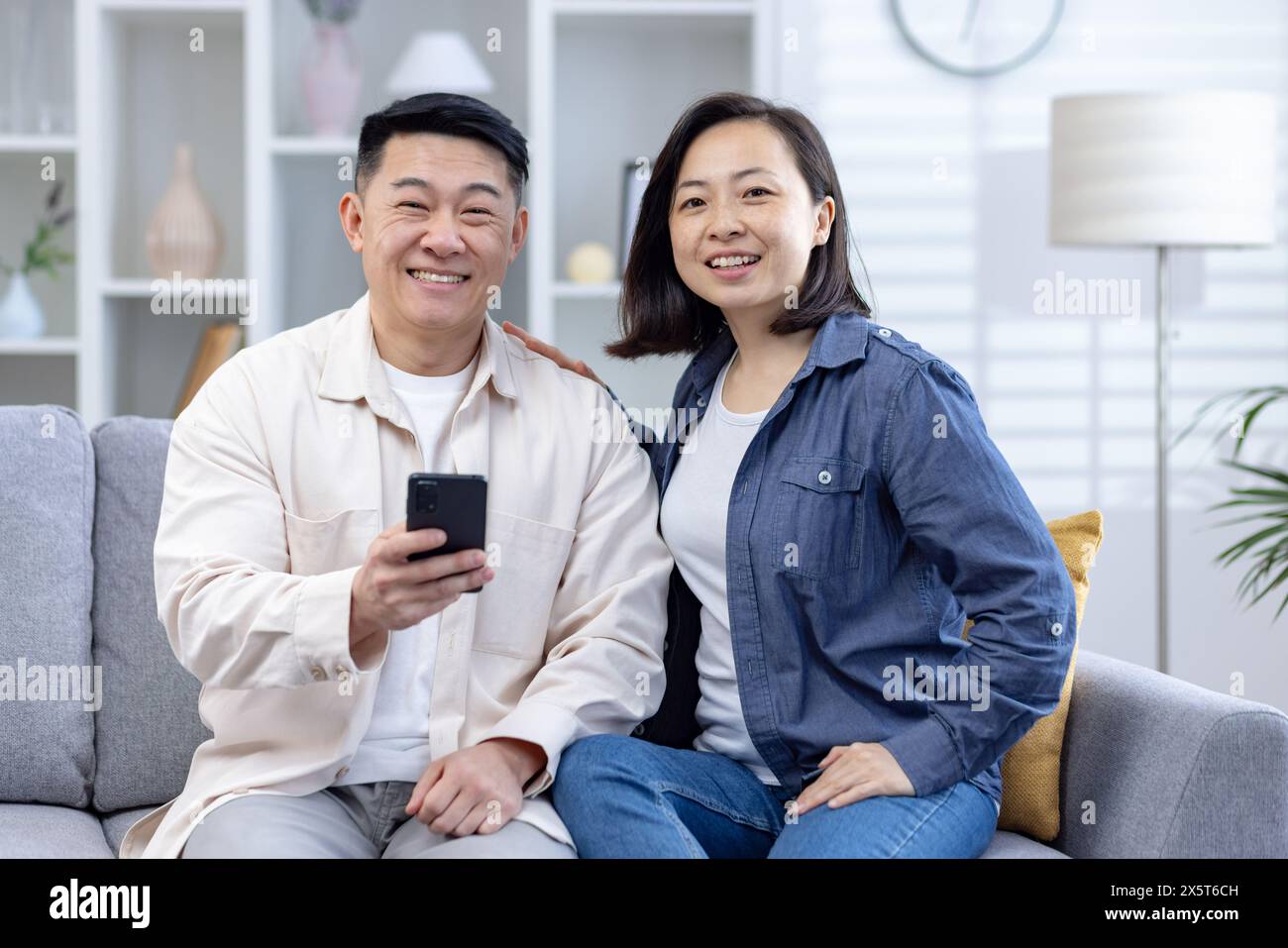 Ein fröhliches Paar mittleren Alters genießt einen entspannten Moment auf dem Sofa im Wohnzimmer. Der Mann hält ein Smartphone, beide lächeln warm. Sie sind lässig gekleidet Stockfoto