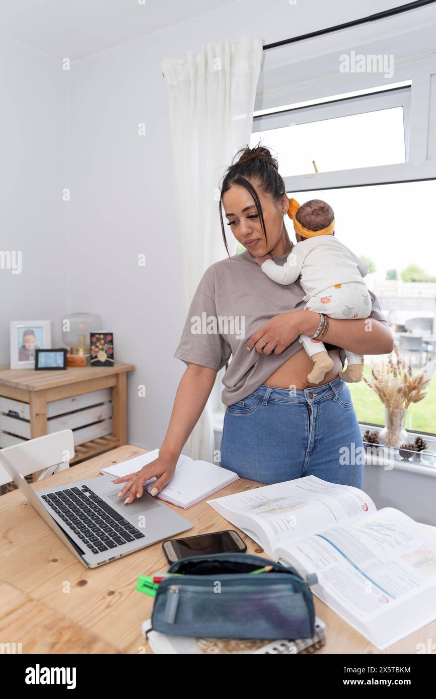 Junge Mutter, die einen Laptop benutzt, während sie ein neugeborenes Mädchen zu Hause trägt Stockfoto