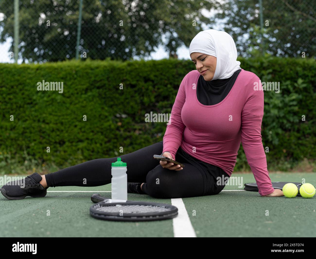 Großbritannien, Sutton, Frau im Kopftuch, die auf dem Tennisplatz sitzt und auf das Smartphone blickt Stockfoto