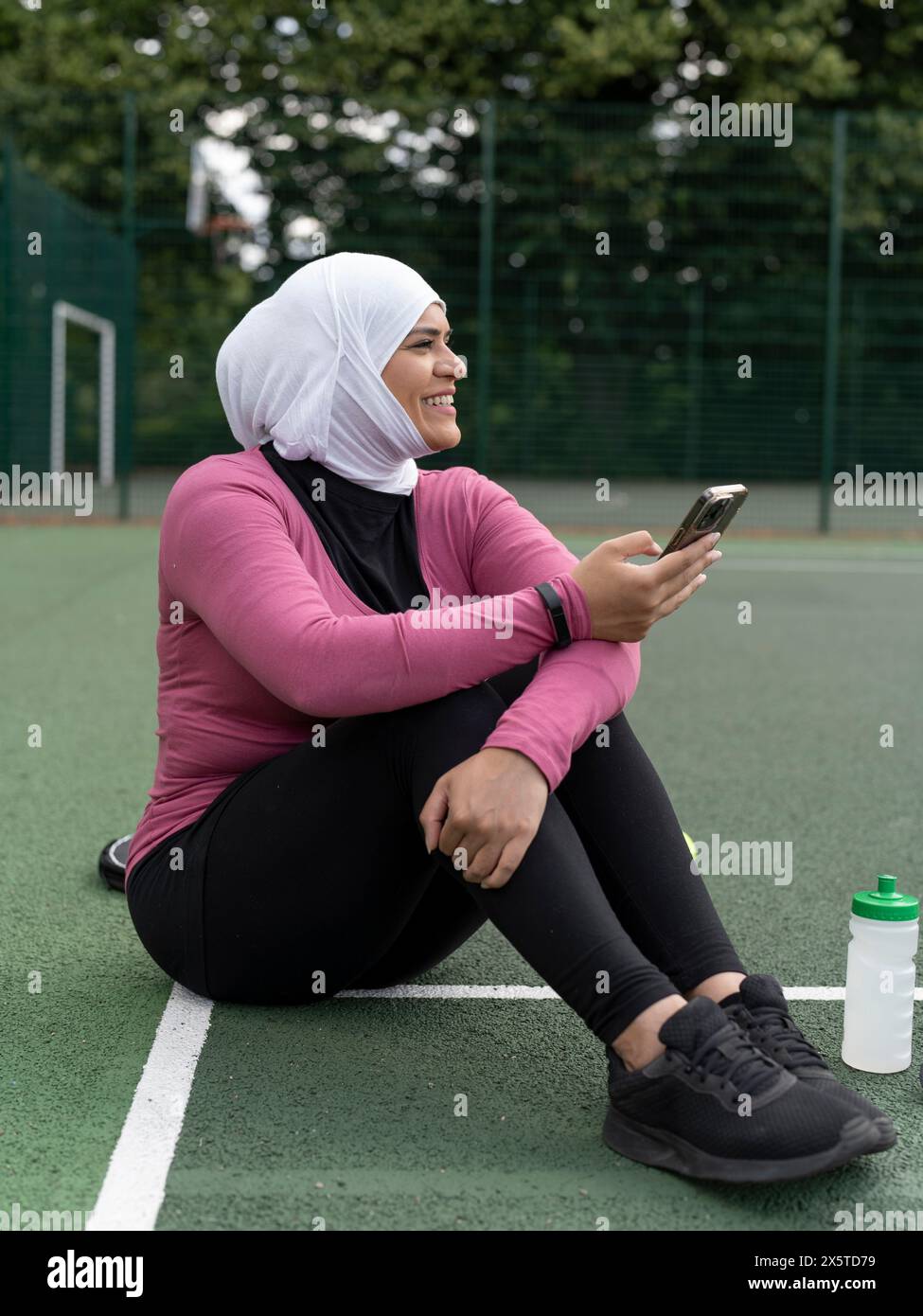 Großbritannien, Sutton, Frau im Kopftuch, die auf dem Tennisplatz sitzt und ihr Smartphone hält Stockfoto