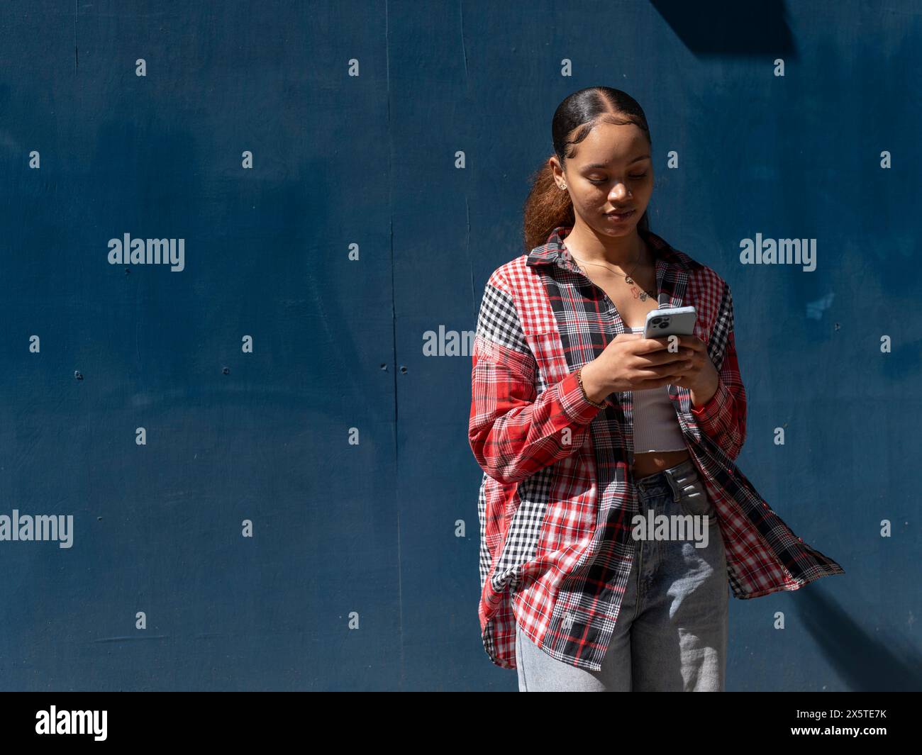 Junge Frau, die mit dem Smartphone an der blauen Wand steht Stockfoto