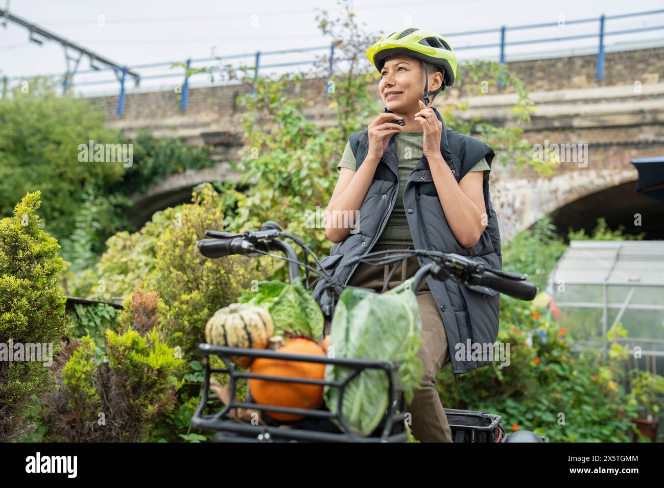 Lächelnde Frau, die den Helm auf einem elektrischen Lastendreirad mit heimischem Gemüse befestigt Stockfoto