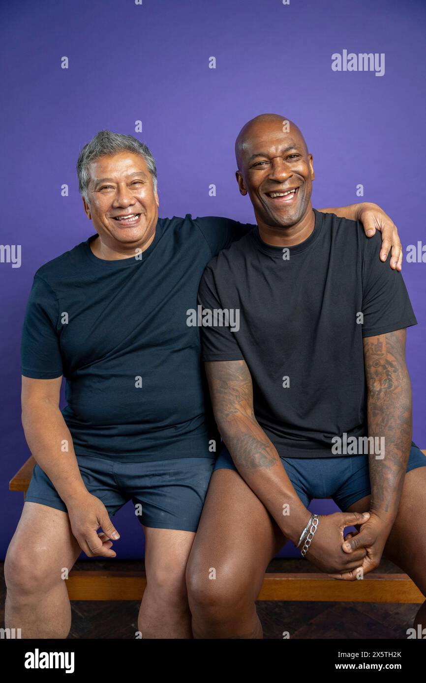 Porträt von zwei lachenden Männern, während sie auf einer Bank sitzen, vor violettem Hintergrund Stockfoto