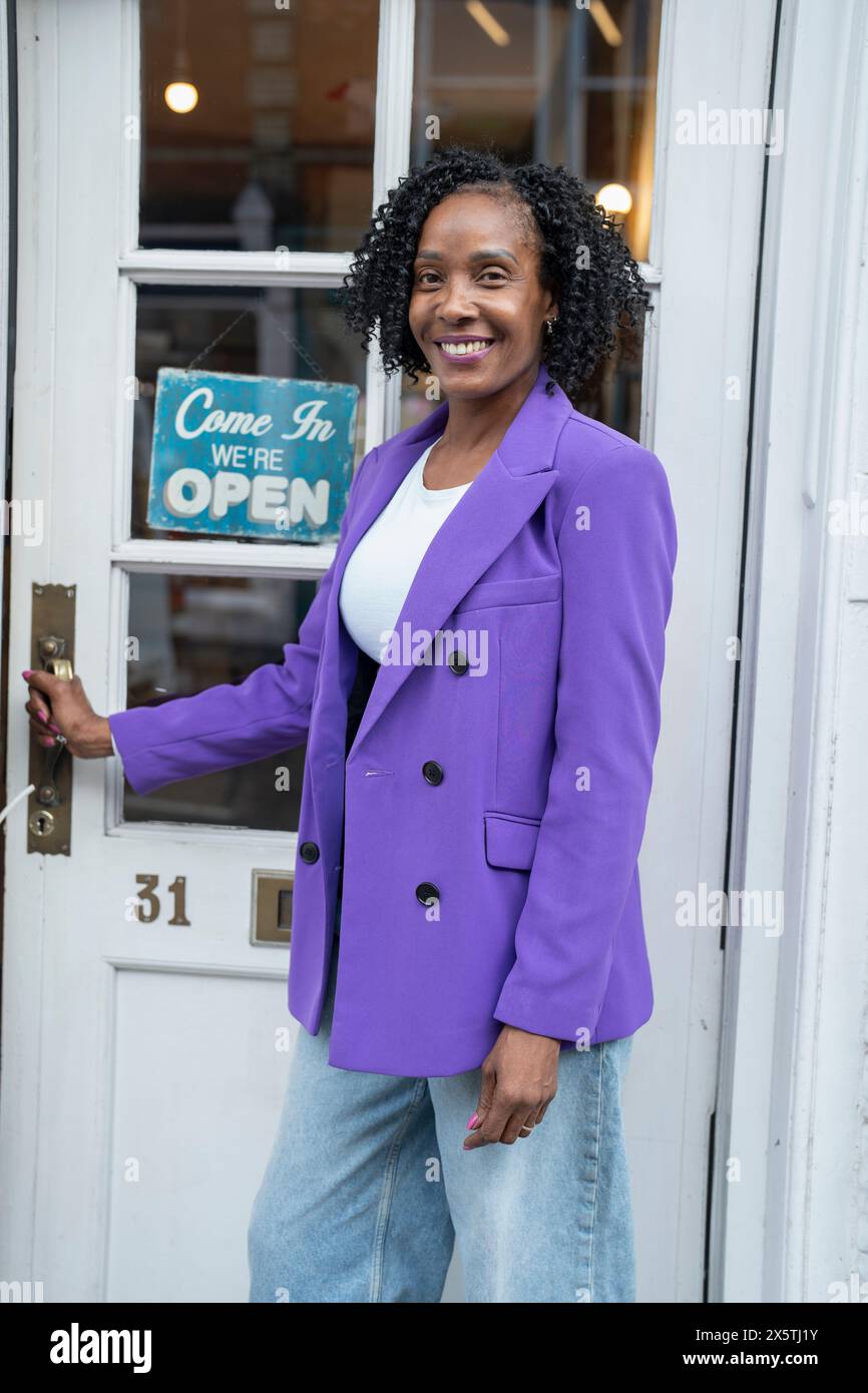 Lächelnde Frau in lila Jacke, die vor einem offenen Laden steht Stockfoto