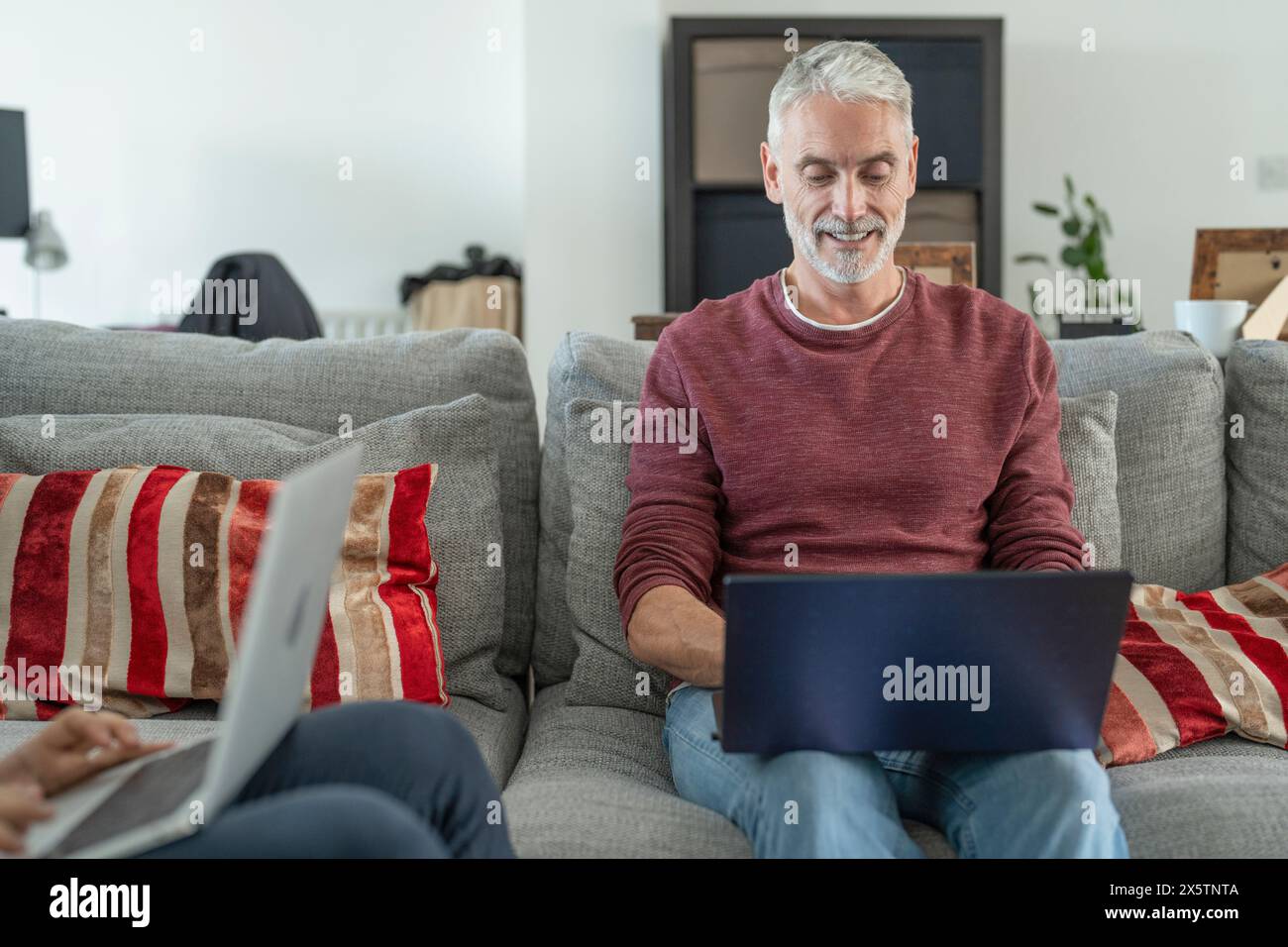 Lächelnder reifer Mann, der zu Hause auf dem Sofa am Laptop arbeitet Stockfoto