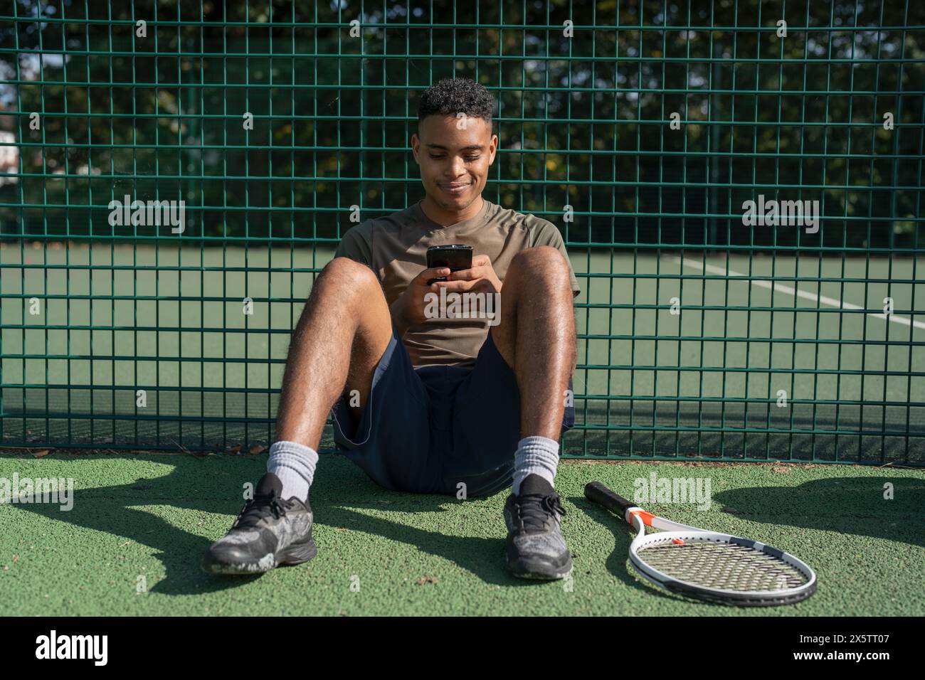 Lächelnder Mann, der auf dem Tennisplatz sitzt und auf das Smartphone schaut Stockfoto