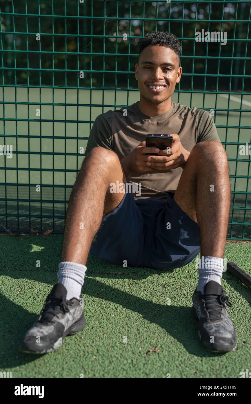 Porträt eines lächelnden Mannes, der auf dem Tennisplatz mit Smartphone sitzt Stockfoto