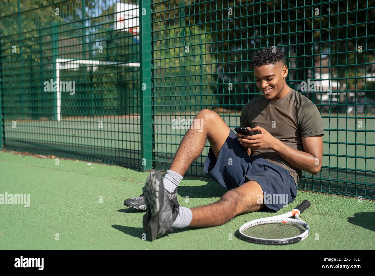 Lächelnder Mann, der auf dem Tennisplatz sitzt und auf das Smartphone schaut Stockfoto