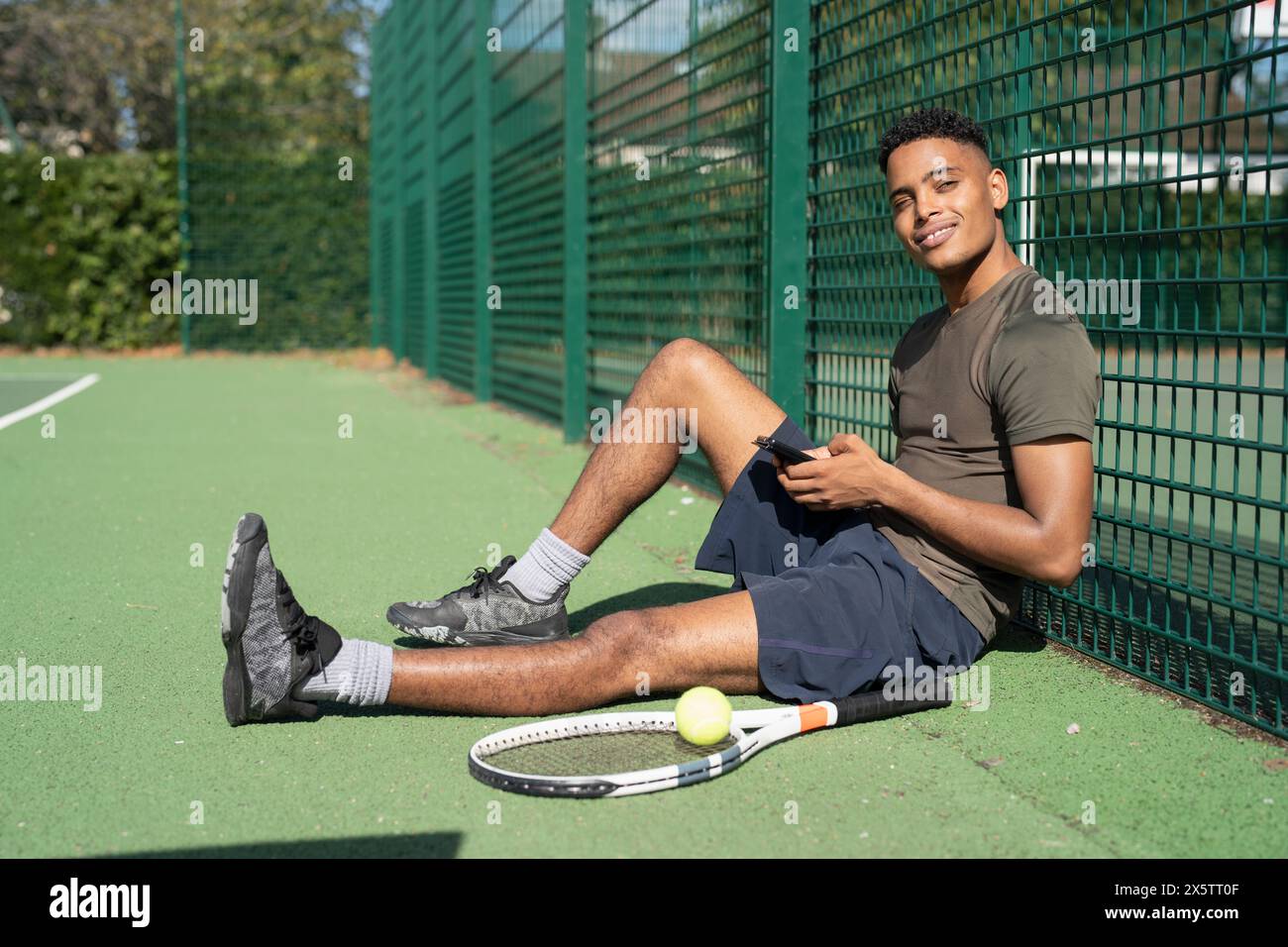 Porträt eines lächelnden Mannes, der auf dem Tennisplatz mit Smartphone sitzt Stockfoto