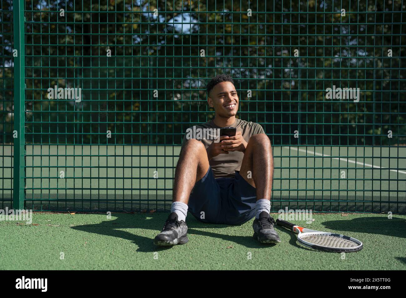 Lächelnder Mann, der auf dem Tennisplatz sitzt und das Smartphone hält Stockfoto