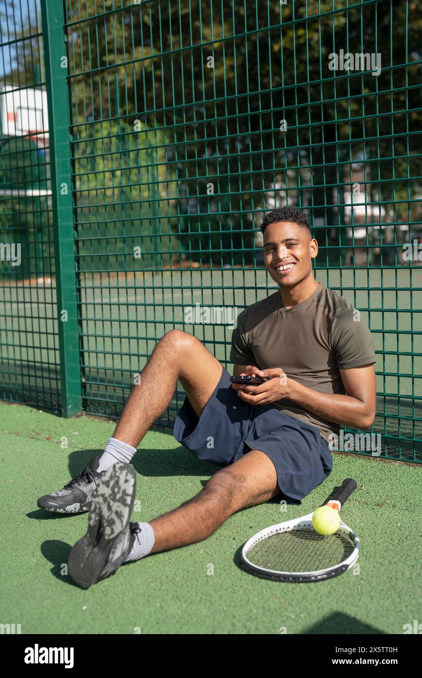 Porträt eines lächelnden Mannes, der auf dem Tennisplatz mit Smartphone sitzt Stockfoto