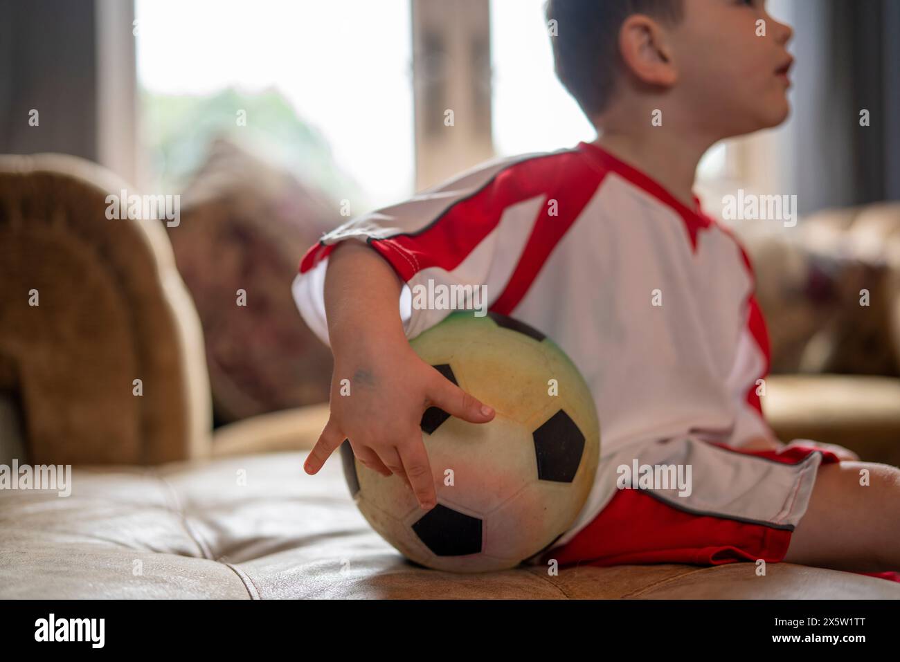 Junge in Fußballuniform sitzt zu Hause mit Ball Stockfoto
