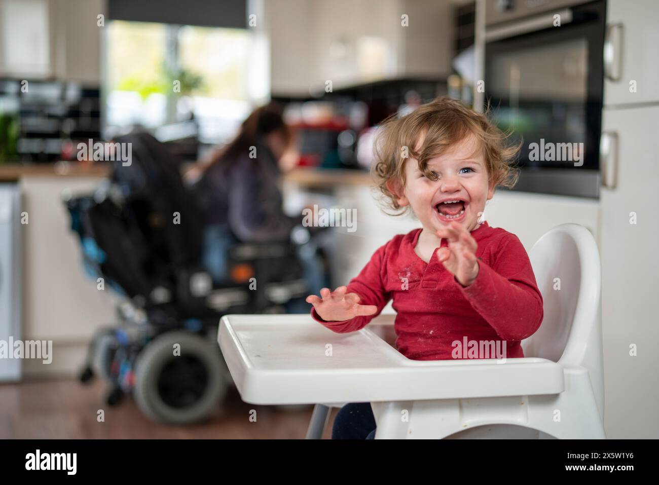 Junge im Hochstuhl mit Mutter im Rollstuhl im Hintergrund Stockfoto