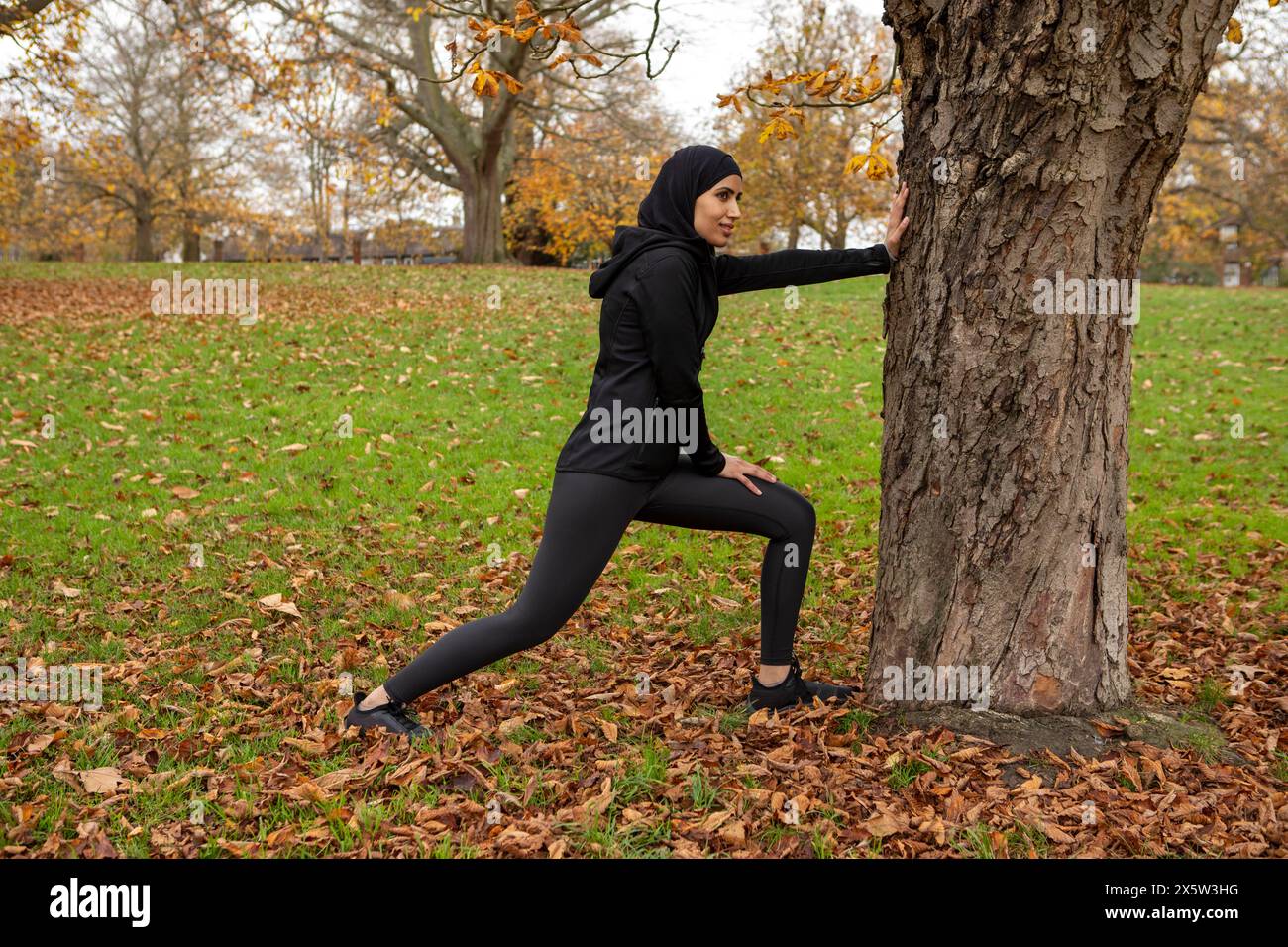 Frau in schwarzer Sportkleidung und Hijab, die sich am Baumstamm im Park ausdehnt Stockfoto