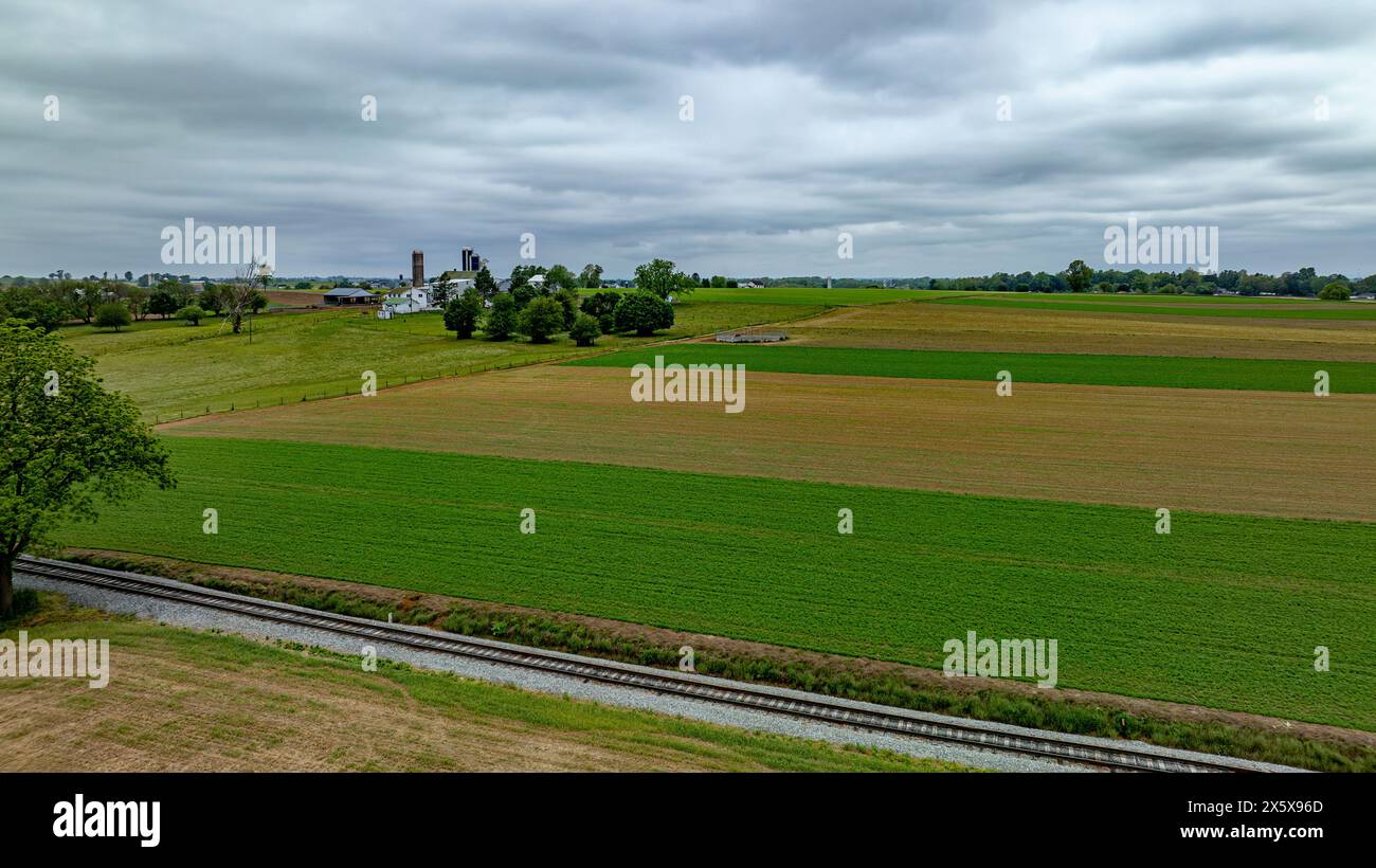 Dieses Luftbild zeigt ein ruhiges Ackerland mit leuchtenden grünen Feldern, die durch die diagonale Weite eines Eisenbahngleises getrennt sind. Die Infrastruktur der Farm, einschließlich Silos und Scheunen Stockfoto