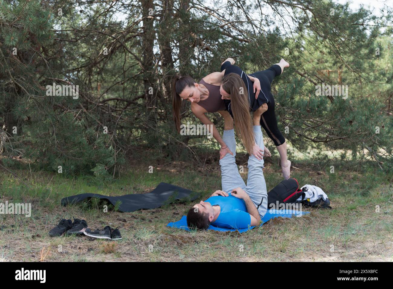 Drei Freunde üben Acro-Yoga-Übungen beim Wandern im Wald. Ein Mann, der auf dem Rücken liegt, hält zwei Mädchen an den Füßen. Stockfoto
