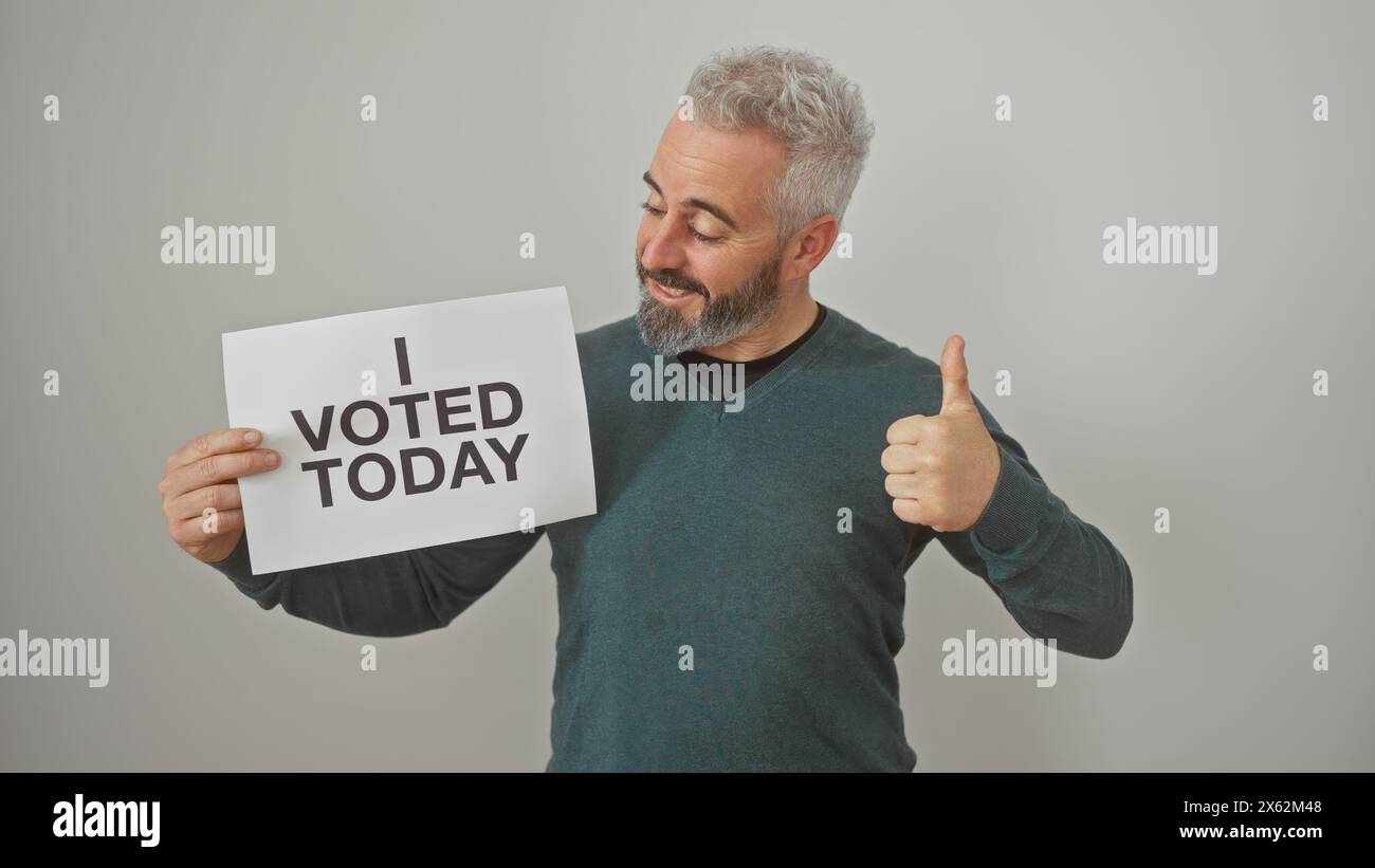 Ein lächelnder bärtiger Mann, der ein Schild mit der Aufschrift "ich habe heute abgestimmt" hält und vor weißem Hintergrund einen Daumen nach oben zeigt Stockfoto