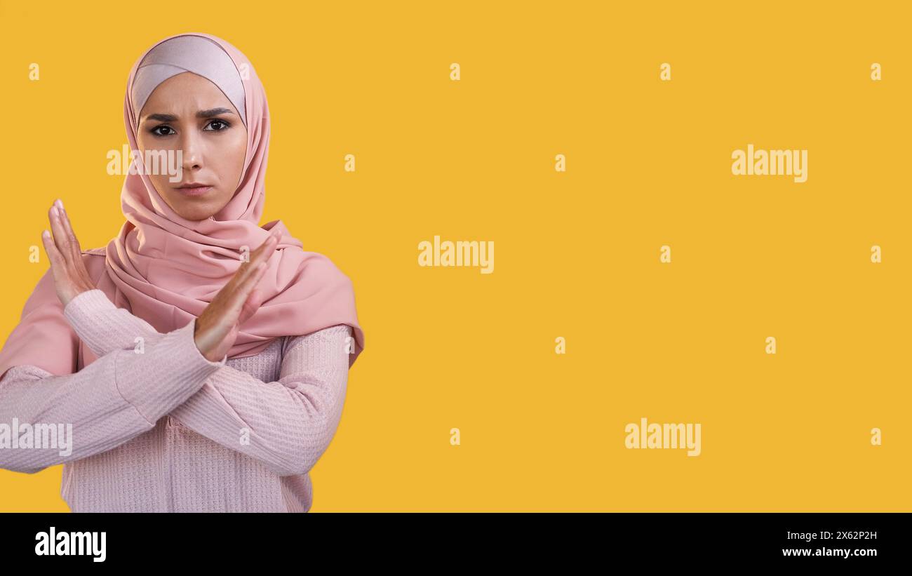 Stoppgeste. Weiblicher Protest. Gestörte strenge Frau im Hijab mit überkreuzten Händen, isoliert auf gelbem Hintergrund. Stockfoto