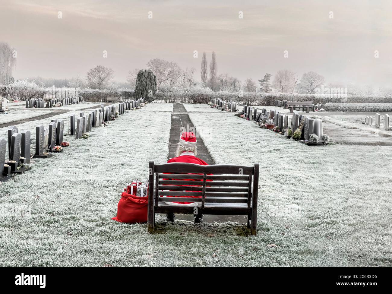 Eine Person, die allein auf einer Holzbank auf einem frostbedeckten Friedhof sitzt, zurück zur Kamera, verkleidet als Weihnachtsmann. UK Stockfoto