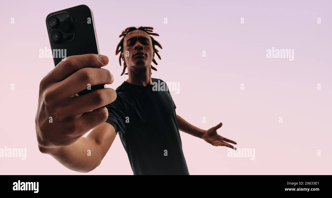 Junger Mann mit Dreadlocks hält ein pinkfarbenes Smartphone und macht mit einer Selfie-Kamera ein Selbstporträt. Gen Z in einem Studiohintergrund, der ihre Verwendung vorstellt Stockfoto
