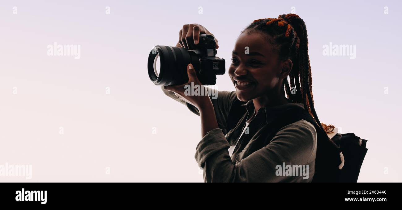 Eine Frau mit Zöpfen hält Momente mit ihrer Digitalkamera fest, während sie im Studio einen Rucksack trägt. Sie verkörpert die Freude der Fotografie und schafft Erinnerungen Stockfoto