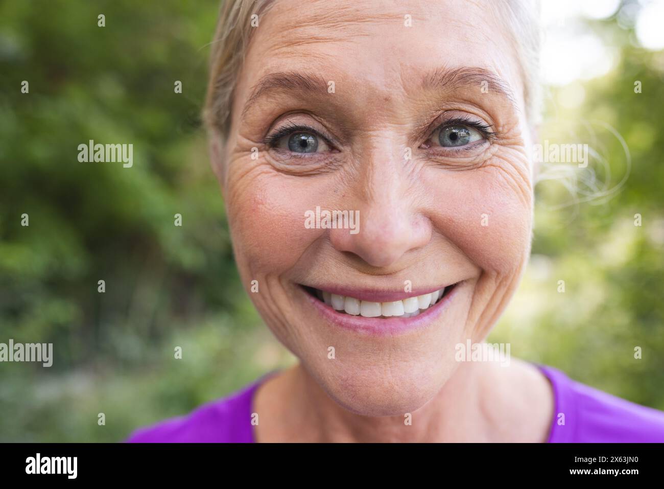 Draußen, eine ältere kaukasische Frau, die ein lila Oberteil trägt und in die Kamera lächelt Stockfoto