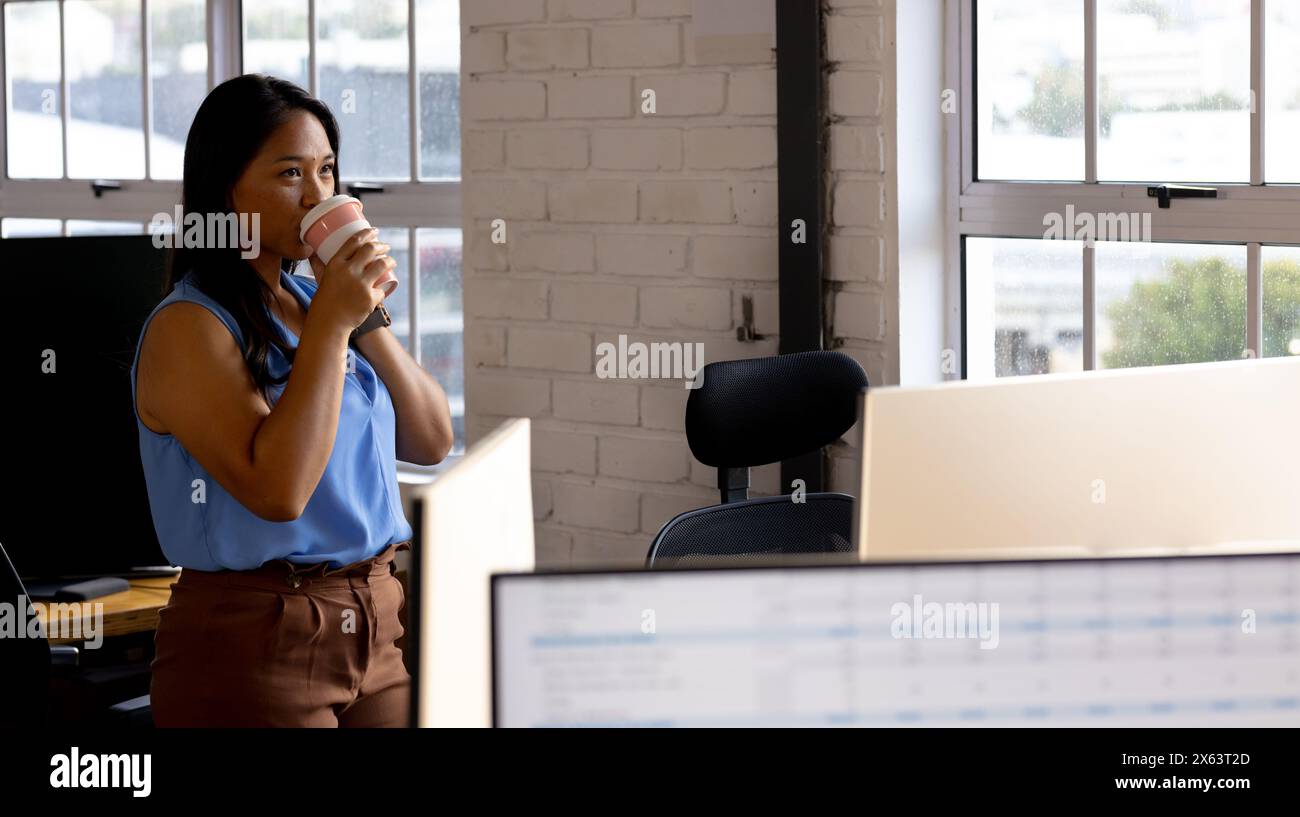 Im Büro trinkt eine Geschäftsfrau mit birassischer Herkunft Kaffee und sieht sich den Computer an Stockfoto
