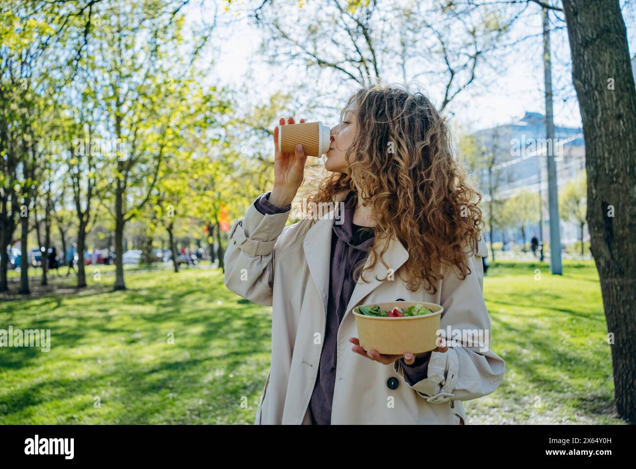 Junge Frau mit lockigen Haaren trinkt Kaffee und hält Salatschüssel, während sie in sonnigem Park steht, gesunder Lebensstil inmitten üppiger Vegetation. Büroangestellte verbringt ihre Mittagspause im Stadtpark. Stockfoto