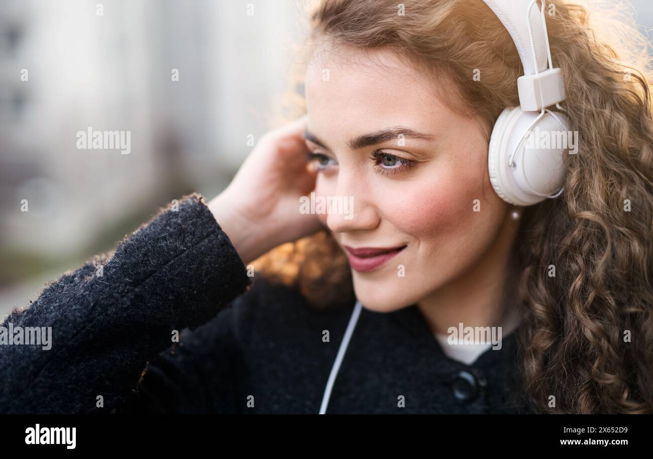 Schöne Frau mit lockigem Haar, die auf der Terrasse steht, den kalten Frühlingsmorgen genießt, lautstarke Musik hört, positive Affirmationen trägt, Kopfhörer trägt. Stockfoto
