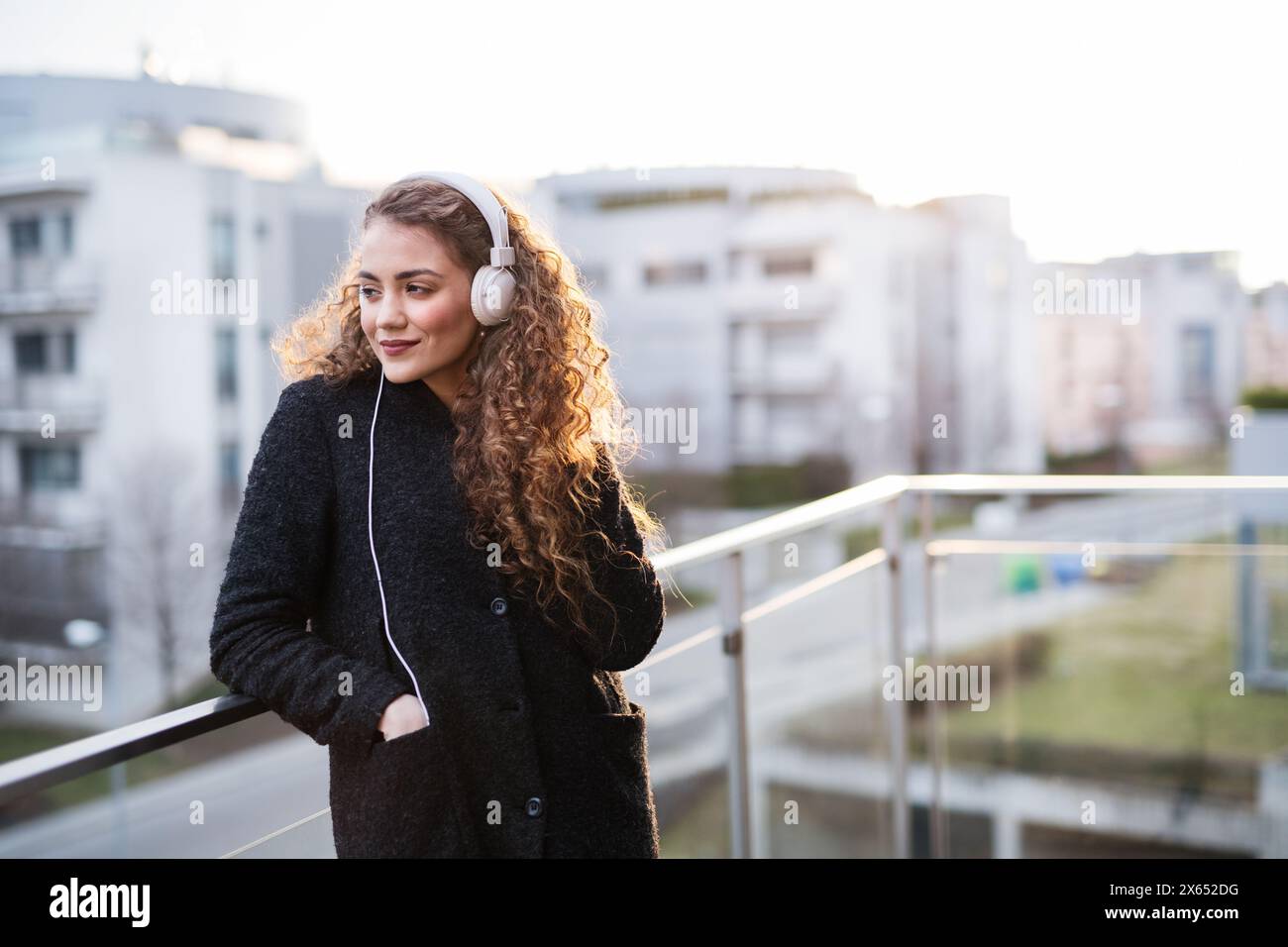 Schöne Frau mit lockigem Haar, die auf der Terrasse steht, den kalten Frühlingsmorgen genießt, lautstarke Musik hört, positive Affirmationen trägt, Kopfhörer trägt. Stockfoto