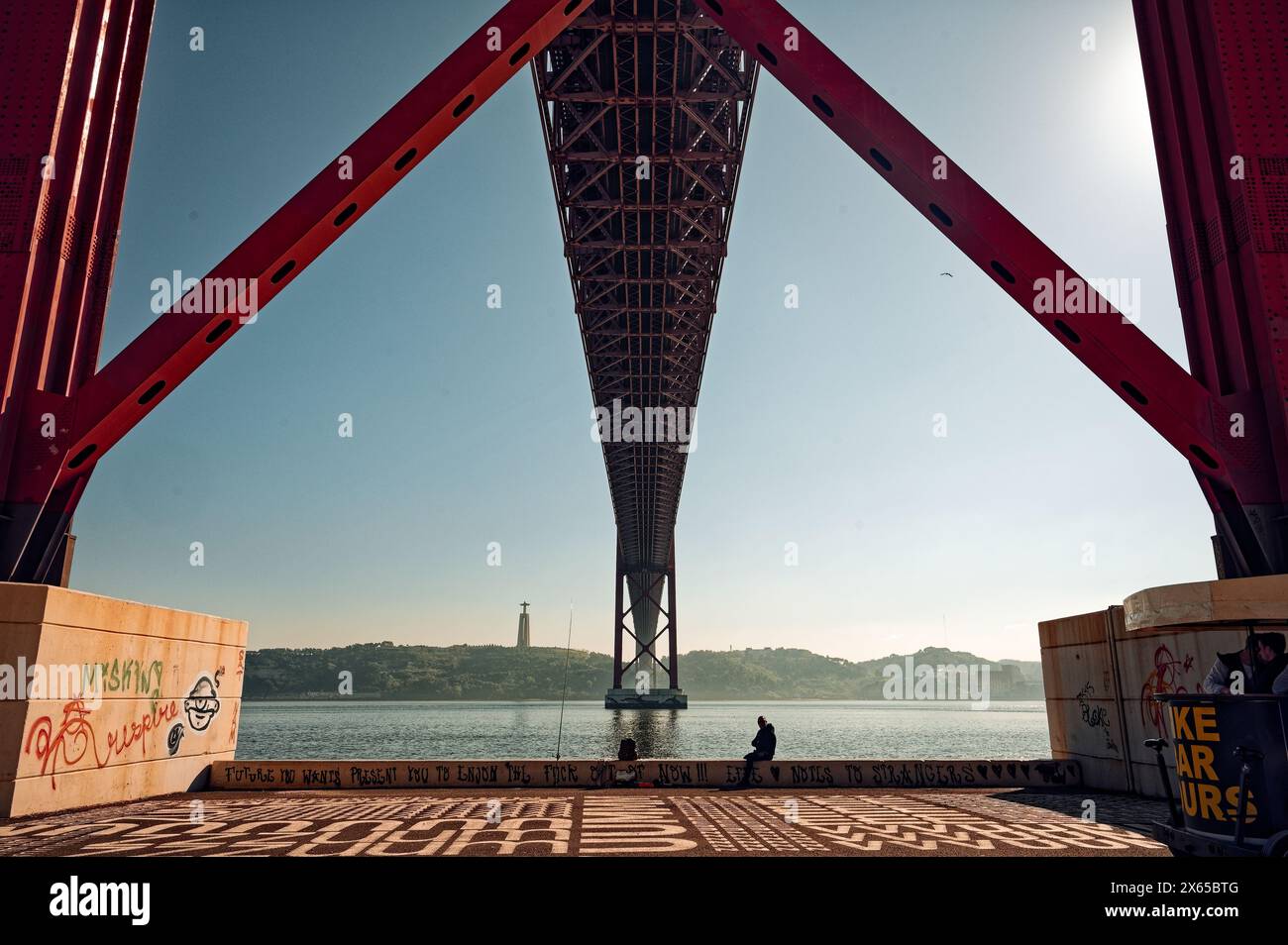 Unter der hoch aufragenden ponte 25 de abril erstreckt sich das Gebäude über den Fluss tejo; eine Einzelfigur ruht am Wasser auf einem mit Graffiti bedeckten Felsvorsprung. Stockfoto