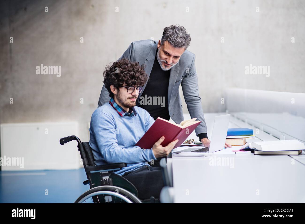 Ein schöner Universitätsprofessor hilft dem Studenten im Rollstuhl und zeigt ihm Buch für seine Diplomarbeit in der Bibliothek. Stockfoto
