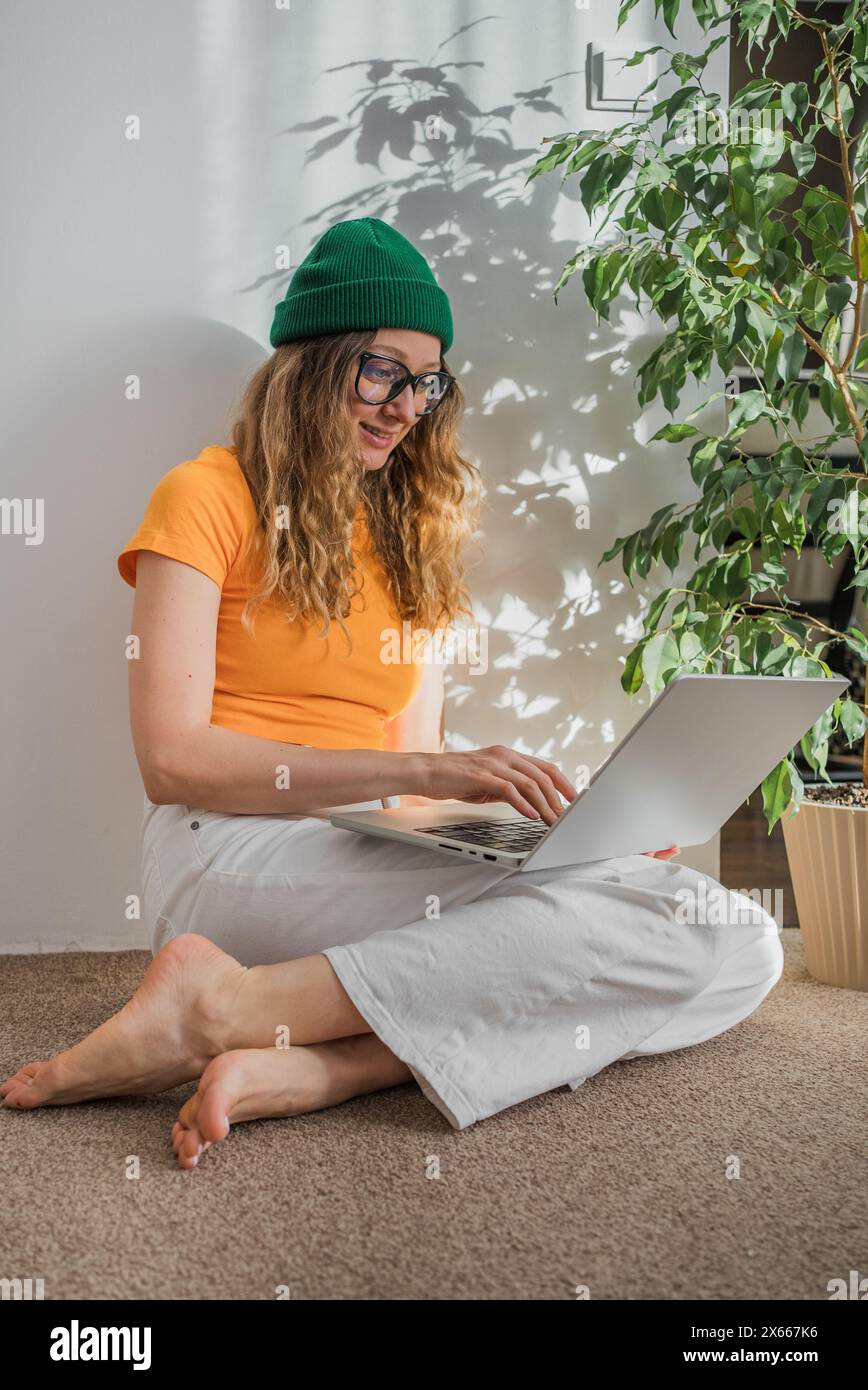 Die lächelnde junge Frau mit lockigem Haar, die eine Brille und eine grüne Mütze trägt, benutzt ihren Laptop bequem in einem sonnendurchfluteten Raum, umgeben von grünen Pflanzen. Stockfoto
