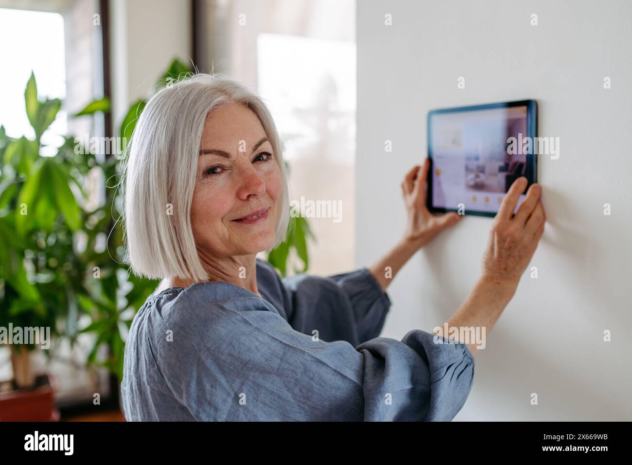 Reife Frau, die den intelligenten Thermostat anpasst, Touchscreen des Smart Home-Geräts. Ältere Frau, die intelligente Technologie zu Hause nutzt. Stockfoto