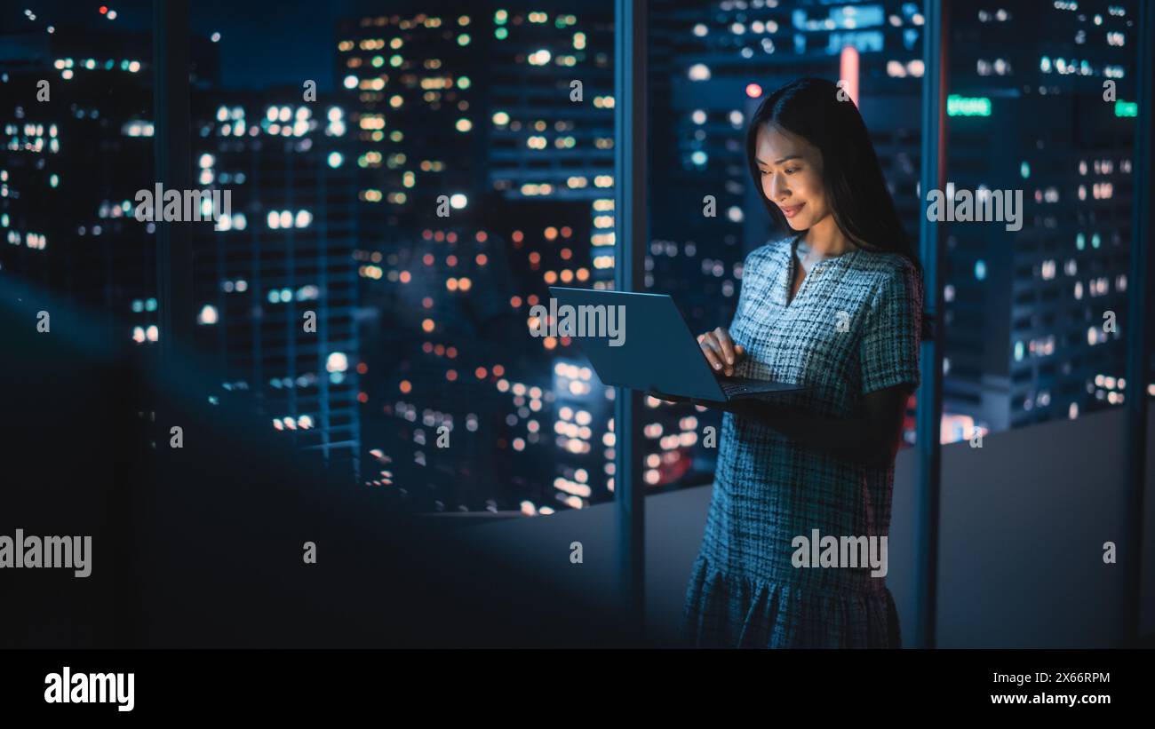 Finanzanalyst mit Laptop, neben dem Fenster stehend mit der wunderschönen Skyline der Nachtsstadt mit Wolkenkratzern. Selbstbewusste asiatische Geschäftsfrau, die in modernen Unternehmensbüros arbeitet. Stockfoto