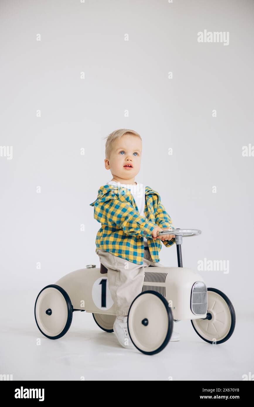 Niedlicher Kleinkinder, der in einem Kinderauto im Zimmer vor weißem Hintergrund spielt und reitet. Glückliche Kindheit, Liebe und Fürsorge. Stockfoto