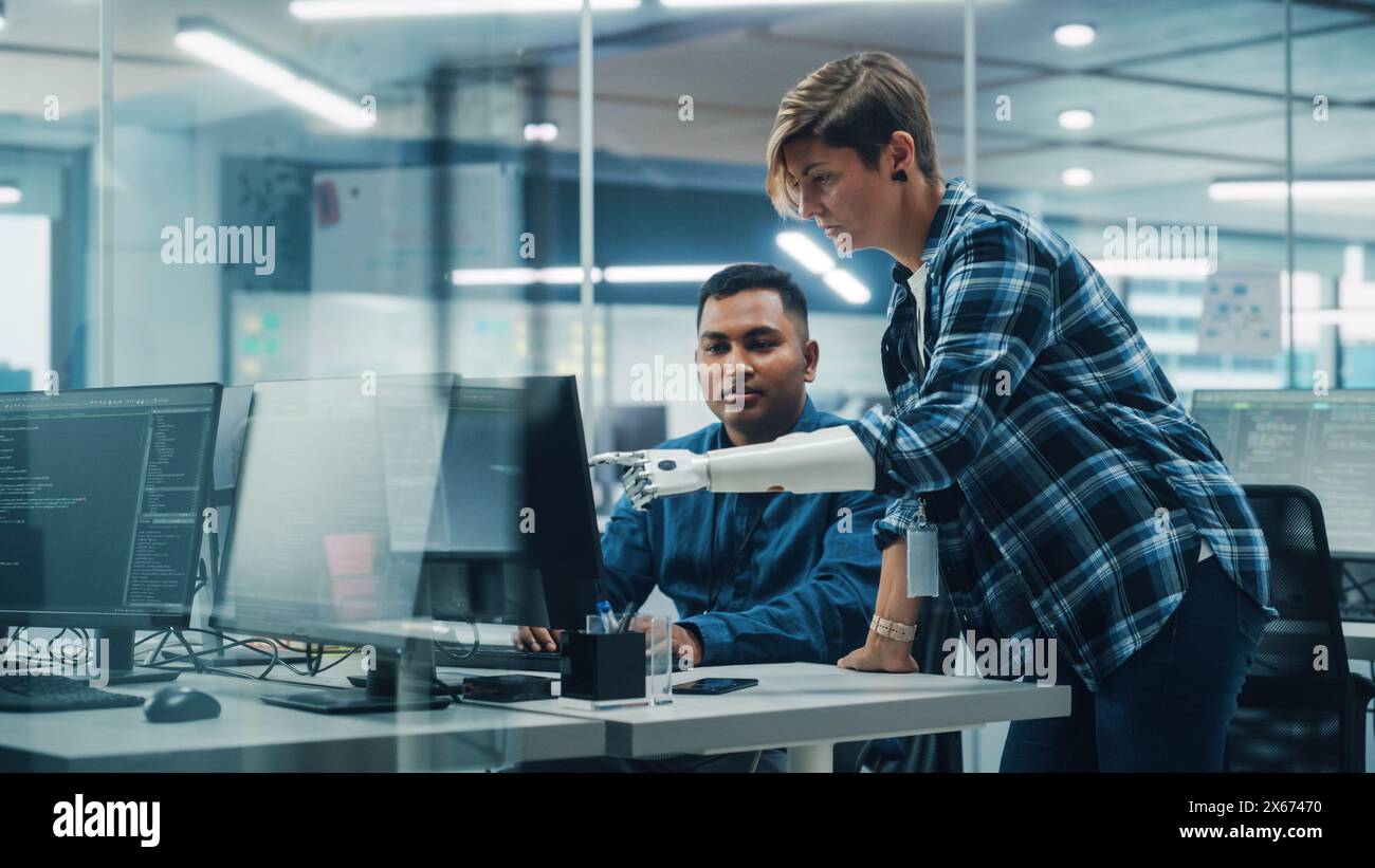 Teamwork in diversen Inclusive Office: Frau mit Behinderung Projektmanager mit Prothetik-Arm spricht mit indischer Fachkraft, die am Desktop-Computer arbeitet. Profis Software Engineers Erstellen App Stockfoto