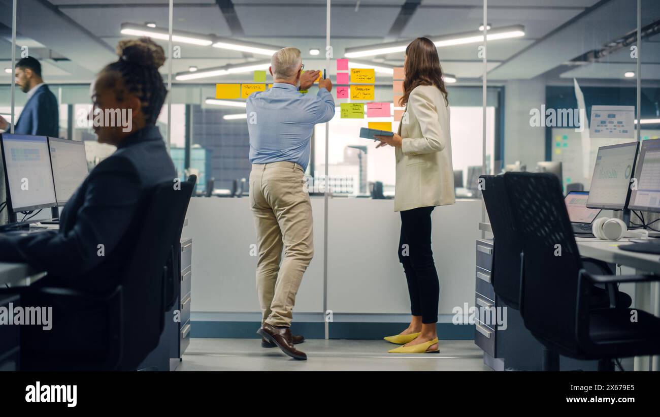 Junger Manager erstellt Projektplan mit einem erfahrenen Senior-Mitarbeiter auf der Bürowand mit Notizen in Papierform. Kollegen diskutieren Geschäfts-, Finanz- und Marketingprojekte. Spezialisten arbeiten in diversen Teams. Stockfoto