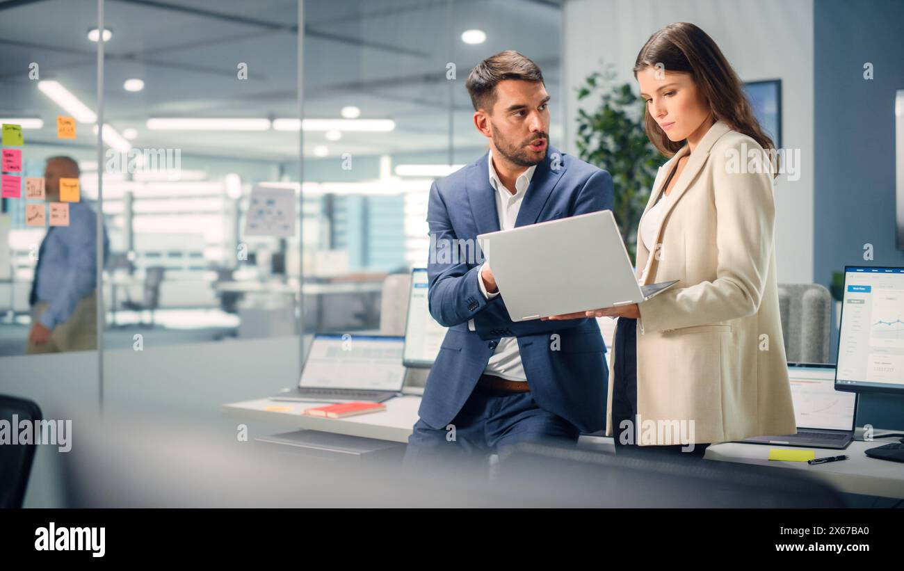 Junger Manager, der mit einem erfahrenen Kollegen spricht, während er im Büro einen Laptop verwendet. Kollegen diskutieren Geschäfts-, Finanz- und Marketingprojekte. Spezialisten arbeiten in diversen Teams. Stockfoto