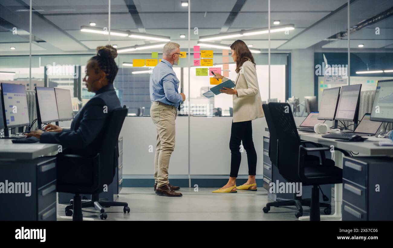 Junger Manager erstellt Projektplan mit einem erfahrenen Senior-Mitarbeiter auf der Bürowand mit Notizen in Papierform. Kollegen diskutieren Geschäfts-, Finanz- und Marketingprojekte. Spezialisten arbeiten in diversen Teams. Stockfoto