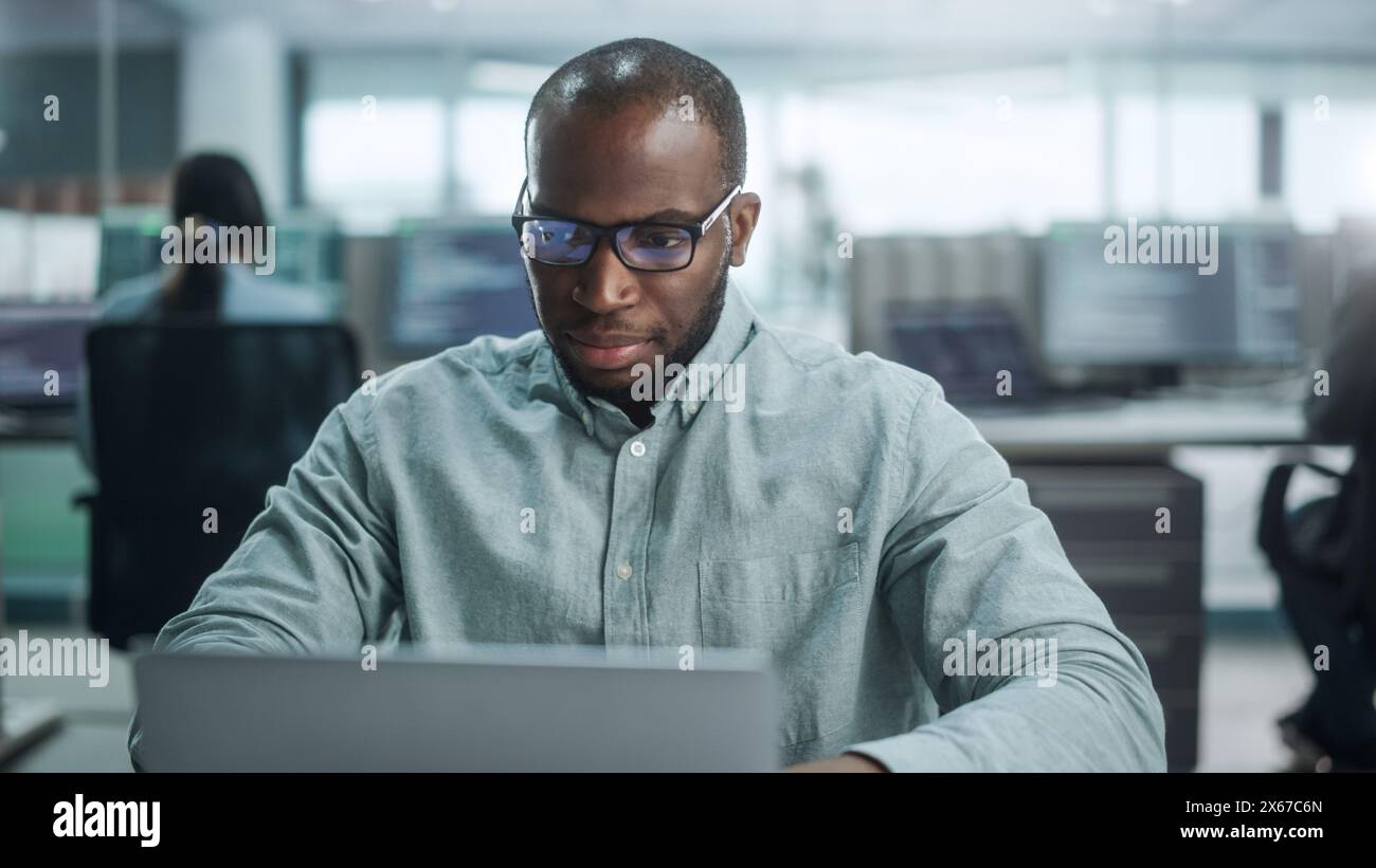 Modernes Büro: Porträt eines durchdachten Black IT Programmierers, der an einem Laptop arbeitet, Lösungen findet, Probleme löst, Brainstorming. Männlicher Software Engineer, Der Brillen Trägt, Entwickelt App, Programm, Videospiel Stockfoto