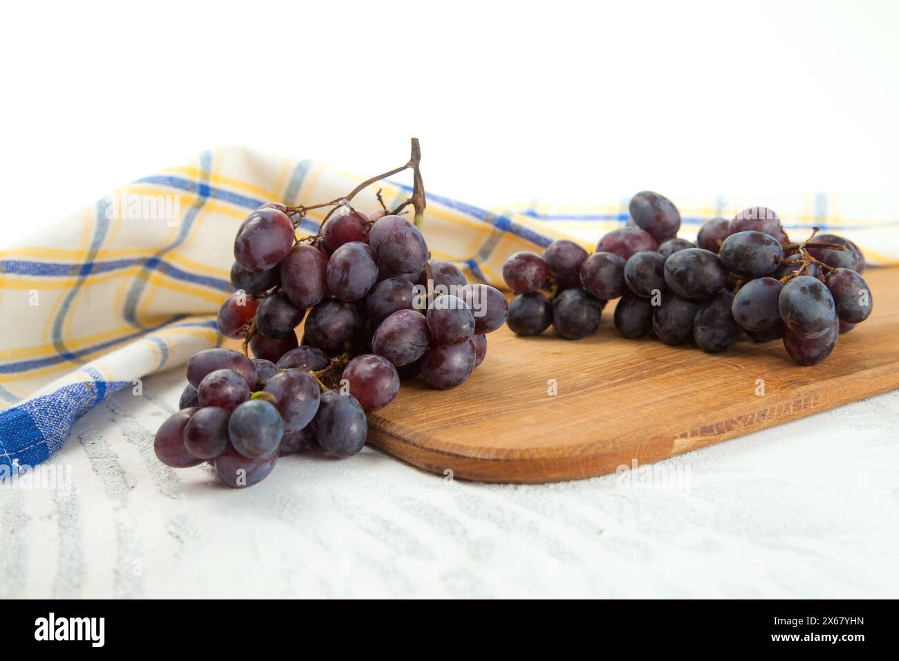 Schneidebrett mit einem großen Haufen reifer dunkelblauer Trauben auf weiß. Lila Weintrauben mit blauem Küchentuch auf weißem hölzernem Hintergrund. Stockfoto